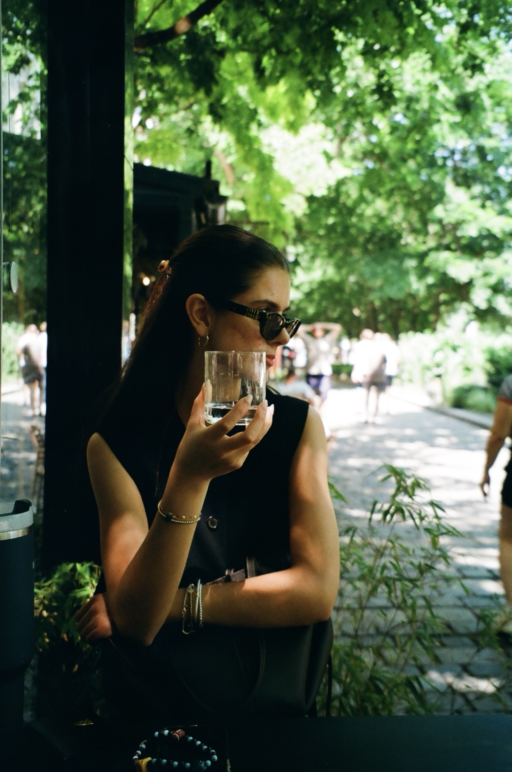 Woman holding a glass beside a sunlit, tree-lined walkway