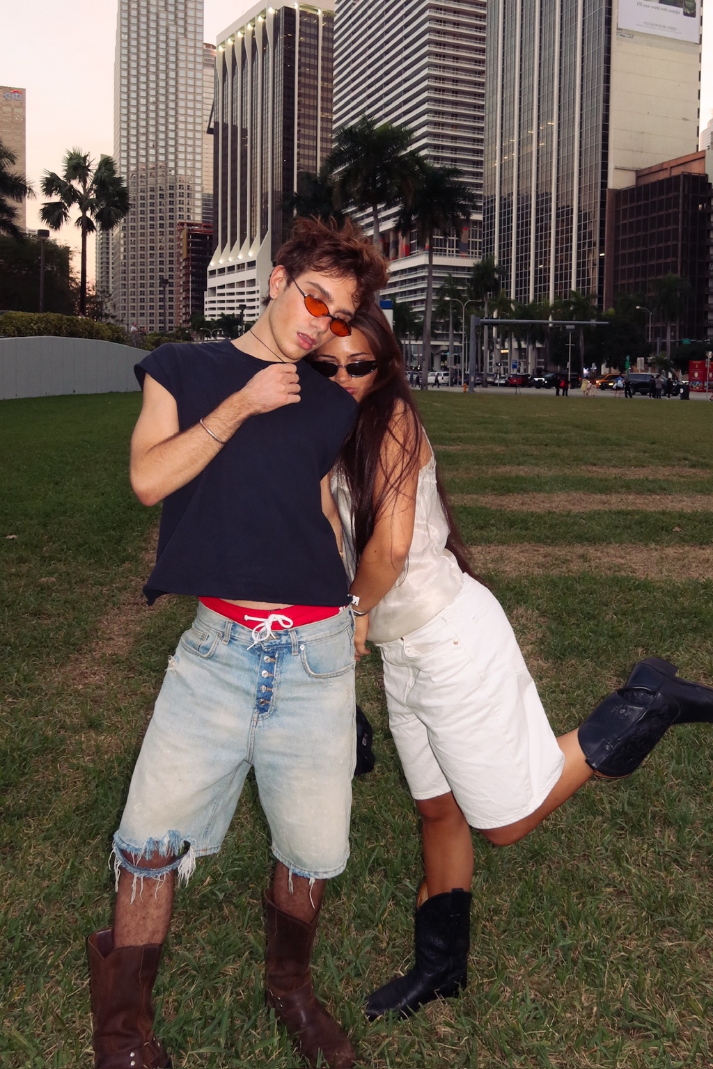 Two people in sunglasses posing on a lawn with palm trees and city buildings
