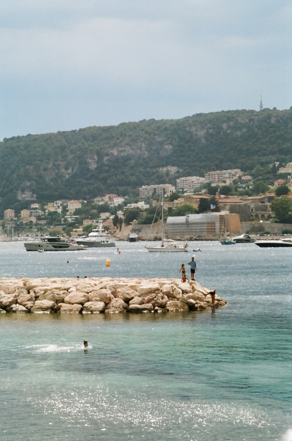 Swimmers beside a rocky breakwater with sailboats in the harbor