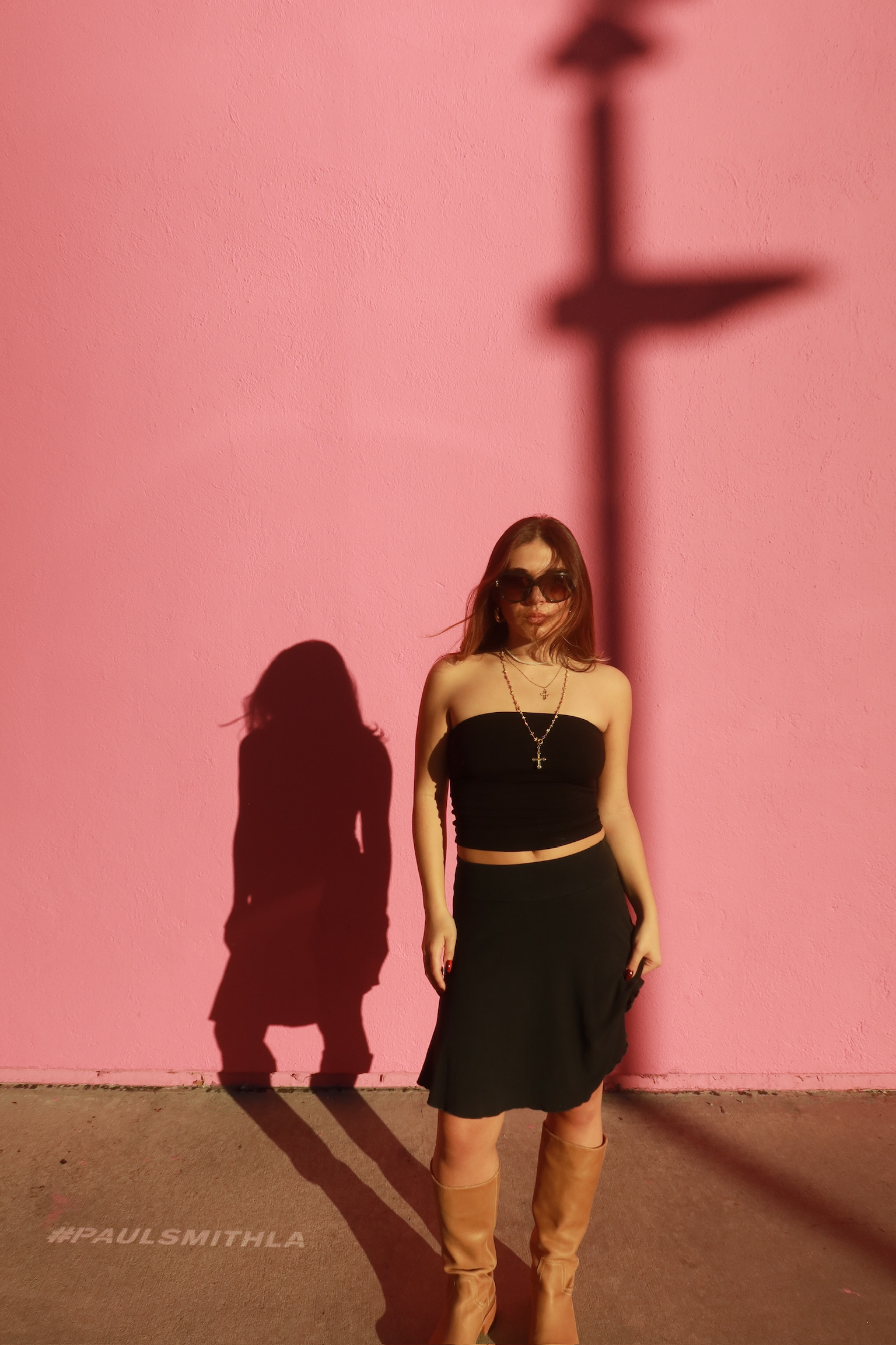 Woman in black clothing and tan boots against a pink wall in sunlight